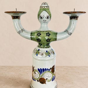 May include: A ceramic candelabra shaped like a stylized figure with a green bodice, a white base featuring blue and brown floral patterns, and two candle holders extending from the arms. The figure has a painted face with dark eyes and a small mouth.