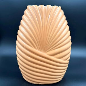 Puede incluir: Un jarrón de cerámica de color beige con un diseño texturizado y acanalado. El jarrón tiene una forma única, con una base ancha que se estrecha hacia la parte superior. El patrón acanalado crea una textura visual, y el jarrón probablemente se utiliza con fines decorativos.