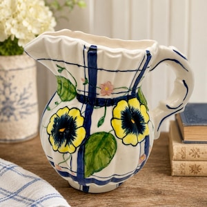 May include: A white ceramic pitcher featuring a scalloped rim, a curved handle, and a hand-painted design of yellow pansies with dark centers, green leaves, and thin blue vertical and horizontal stripes forming a grid pattern across the surface.