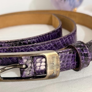 May include: A purple snakeskin-patterned belt with a silver-tone buckle. The belt is thin and appears to be made of leather. The buckle is rectangular with the brand name "miu miu" engraved on it.