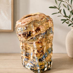 May include: A hand-built ceramic vase with an irregular, textured surface featuring a glossy glaze in shades of amber, brown, and deep blue. The vessel has a wide, uneven opening at the top and a blocky, organic shape with visible indentations.