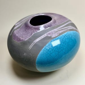 May include: A round ceramic vase with a matte charcoal gray base. The surface features glossy, crackled glaze sections in vibrant turquoise and soft purple, creating a distinct contrast against the dark, textured, unglazed gray body.