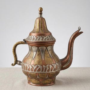 May include: An antique copper teapot with intricate metalwork. The teapot features a long spout, a curved handle, and a decorative lid. The body of the teapot is adorned with patterns in silver and gold tones, creating a vintage aesthetic.