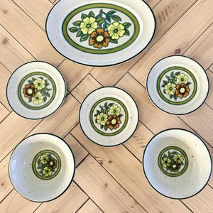 Könnte beinhalten: Ein Vintage-Keramik-Geschirrset mit einer ovalen Platte, vier kleinen Tellern und zwei Schalen. Jedes Stück hat einen weißen, gesprenkelten Hintergrund, ein grünes und orangefarbenes Blumendesign und einen schwarzen Rand. Das Set ist auf einer hellen Holzoberfläche angeordnet.
