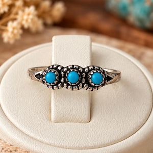 May include: A silver-toned metal ring featuring three small, round, bright blue turquoise-coloured stones set in individual scalloped bezels. The band has a textured, oxidised finish with decorative triangular cutouts on each side.