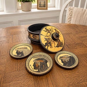 Op de afbeelding: Een decoratieve zwart-gouden doos met een deksel met een geschilderde scène van figuren in actie. Drie bijpassende onderzetters met portretten in Griekse stijl omringen de doos. De onderzetters en de doos hebben een goud- en zwartkleurenschema.