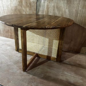 Puede incluir: Mesa auxiliar ovalada de madera con acabado oscuro. La tapa está soportada por un marco con paneles de mimbre tejido y una distintiva pata en forma de V. La mesa es de color marrón cálido y tiene veta natural de madera.