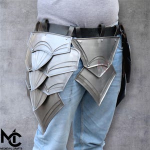 May include: Silver-coloured medieval armour leg protection, featuring overlapping metal plates. The armour is attached to a black belt and worn over blue jeans. The image includes the Medieval Crafts logo.