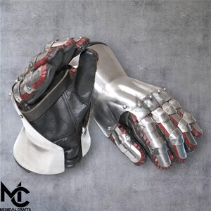 May include: A pair of medieval gauntlets. The gauntlets are made of polished metal plates and black leather. The interior of the gauntlets is red. The gauntlets are designed to protect the hands and wrists.