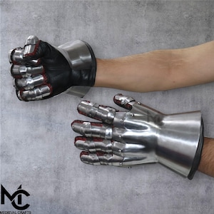 May include: Pair of steel gauntlets with articulated fingers and a flared cuff. The gauntlets have a polished metal finish, black leather palms, and red fabric lining the finger joints. The Medieval Crafts logo is visible in the lower left corner.