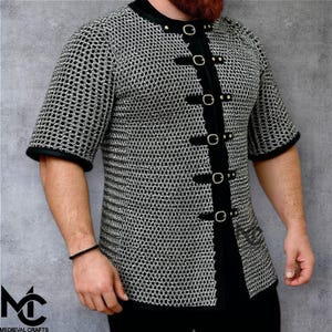 May include: Short-sleeved chainmail shirt with black trim and a black front panel secured by gold-coloured buckles. The shirt is made of metal rings and is designed for historical reenactment or costume use.