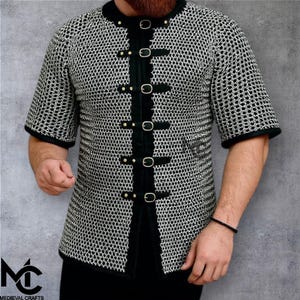 May include: Short-sleeved chainmail shirt with black trim and buckle fastenings. The shirt has a round collar and is made of metal rings. The image includes the logo "Medieval Crafts" in the lower left corner.