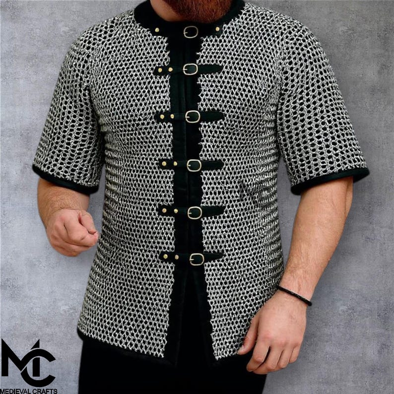 May include: Short-sleeved chainmail shirt with black trim and buckle fastenings. The shirt has a round collar and is made of metal rings. The image includes the logo "Medieval Crafts" in the lower left corner.