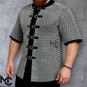 May include: A short-sleeved chainmail shirt with black trim and fasteners. The shirt has a black collar and is fastened with black straps and silver buckles. The image includes the logo "MEDIEVAL CRAFTS" in the bottom left corner.