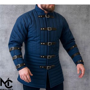 May include: A blue quilted jacket with a stand-up collar and black leather straps with buckles. The jacket has horizontal and vertical stitching, and the sleeves also feature black leather straps. The logo "Medieval Crafts" is visible in the bottom left corner.