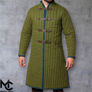 Può includere: Gambesone medievale trapuntato verde oliva con collo alto e maniche lunghe. L'indumento presenta tre cinturini in pelle marrone con fibbie sul davanti e finiture blu. Il logo Medieval Crafts è visibile in basso a sinistra.