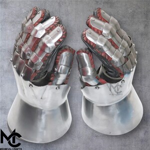 May include: A pair of medieval gauntlets, crafted from polished steel plates, red leather, and black fabric. The gauntlets feature articulated fingers and a flared cuff. The logo "MEDIEVAL CRAFTS" is visible. Suitable for historical reenactment or display, these gauntlets are a striking example of historical armour.