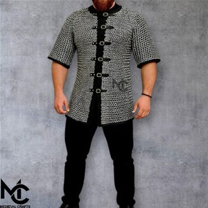 May include: A short-sleeved chainmail shirt with black leather trim and fasteners. The shirt is made of small, interlocking metal rings and features a high collar. The shirt is worn with black trousers. The Medieval Crafts logo is visible.