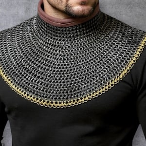 Puede incluir: Un cuello de cota de malla plateado con un ribete dorado, usado sobre una camisa negra de manga larga. La cota de malla está hecha de pequeños anillos de metal entrelazados. El cuello tiene una banda de cuero marrón.