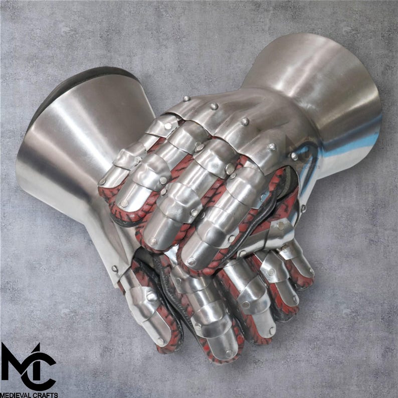 May include: A pair of medieval gauntlets made from polished steel, with articulated finger plates and a red interior. These gauntlets are designed to protect the hands and wrists. The image shows the gauntlets against a grey background. The logo "Medieval Crafts" is visible.