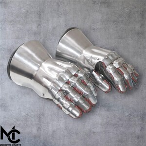May include: A pair of polished steel gauntlets, with articulated finger plates and riveted construction. The interior of the fingers is lined with red fabric. The gauntlets have a flared cuff and are designed for protection. The logo "MEDIEVAL CRAFTS" is visible.