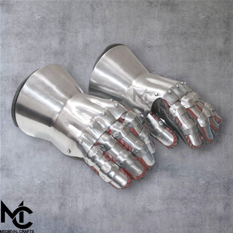 May include: A pair of polished steel gauntlets, with articulated finger plates and riveted construction. The interior of the fingers is lined with red fabric. The gauntlets have a flared cuff and are designed for protection. The logo "MEDIEVAL CRAFTS" is visible.