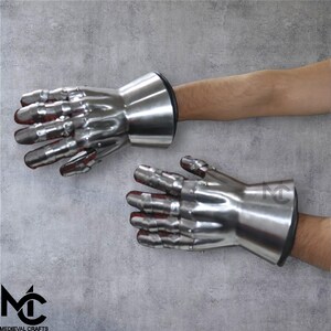 May include: Pair of polished steel gauntlets with articulated fingers and flared cuffs. The interior of the fingers is red. These gauntlets are designed to protect the hands and wrists. The Medieval Crafts logo is visible in the lower left corner.