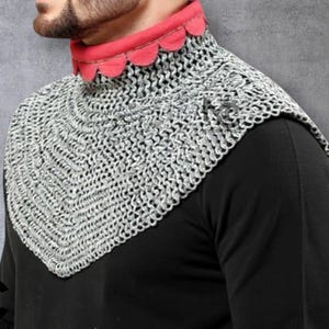 May include: A silver chainmail coif with a red scalloped collar. The chainmail is made of small, interlocking metal rings. The coif is worn over a black garment. The Medieval Crafts logo is visible in the lower left corner.