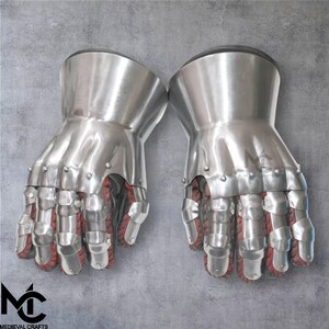 May include: Pair of polished steel gauntlets with articulated fingers and red interior lining. The gauntlets have a flared cuff and are riveted together. The Medieval Crafts logo is visible in the lower left corner. These gloves are designed for historical reenactment or display.