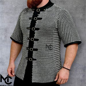 May include: A short-sleeved chainmail shirt with black trim and a black front closure. The shirt has a high collar and is fastened with metal clasps. The logo "MEDIEVAL CRAFTS" is visible in the lower left corner.