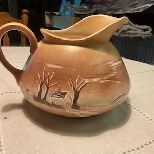 May include: A vintage ceramic pitcher with a tan and brown landscape design. The pitcher has a large handle and a spout. The landscape features trees and a small house. A decorative piece for the home.