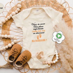May include: Cream-colored baby onesie with the text "Veux-tu avoir Une place importante DANS MA VIE en devenant MA MARRAINE?" in black and orange. The onesie is paired with brown baby shoes, a pacifier, and wooden beads.