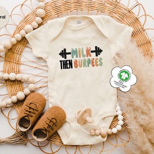 May include: Cream-colored baby onesie with the text "MILK THEN BURPEES" in black, green, and orange. The onesie features a barbell graphic. The image includes brown baby shoes, a pacifier, and decorative wooden beads. The onesie is made of organic textile.