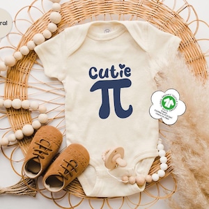 May include: A cream-colored baby onesie with the words "Cutie π" in navy blue. The onesie is laid on a woven wicker surface with a pair of brown baby shoes, a pacifier, and decorative wooden beads. A natural-colored label is in the upper left corner.