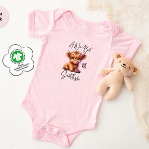 May include: A light pink baby onesie with the text "A Wee Bit Scottish" and an illustration of a Highland cow. A small stuffed bear and a decorative cloth are next to the onesie. The onesie has short sleeves and snaps at the bottom.