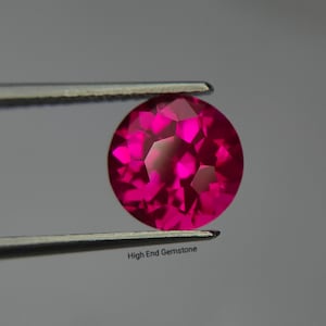 May include: A round, faceted, vibrant pinkish-red gemstone held by metal tweezers. The stone displays a brilliant cut with multiple reflective surfaces. Below the gemstone, the text "High End Gemstone" is printed in a small, clear black font.
