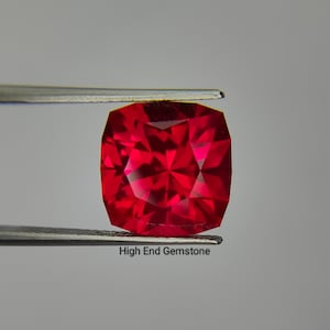 May include: A vibrant red cushion-cut gemstone held by silver-toned metal tweezers. The gemstone features a faceted surface that reflects light, creating a deep crimson appearance. The text High End Gemstone is printed below the gemstone.