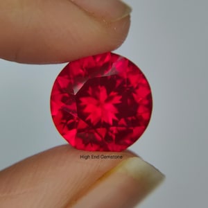 May include: A faceted, round, bright red gemstone. The gem is held between two fingers against a light background. The text "High End Gemstone" is visible at the bottom of the image.