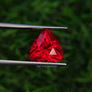 May include: A vibrant red gemstone cut into a triangular shape with multiple facets that reflect light. The gemstone is held between two thin metal prongs. The text High End Gemstone is printed in small white letters at the bottom of the image.