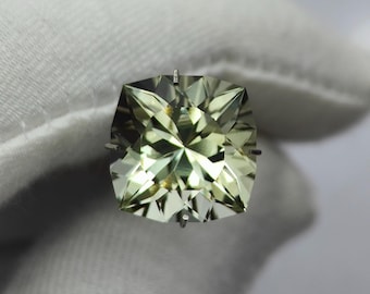 May include: A faceted, square-cut gemstone with a pale green hue. The gem is held by a silver setting, and the light reflects off the many facets. The gemstone is displayed against a white background.