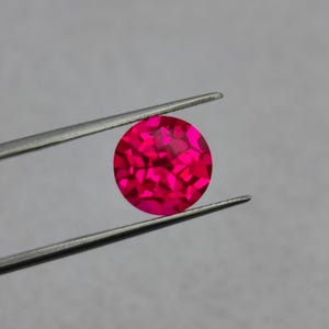May include: A round, faceted, hot pink gemstone is held by silver tweezers against a grey background. The gemstone's surface reflects light, creating a sparkling effect. The gem is a vibrant, eye-catching focal point.