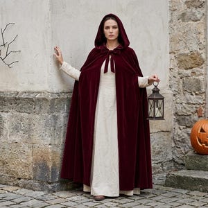May include: A floor-length, deep burgundy velvet hooded cloak with a front tie closure, worn over a long-sleeved, cream-colored linen-style dress. The cloak features a structured hood and a smooth, soft-touch fabric finish that drapes to the floor.