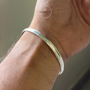 May include: A smooth, polished silver-toned bangle bracelet with a rounded, solid profile. The metal surface reflects light, creating bright highlights along the curve of the band. The word SURETRAM is visible in a small circular logo overlay.