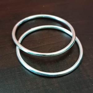 Puede incluir: Dos brazaletes plateados apilados sobre una superficie marrón oscuro. Los brazaletes son circulares y tienen una superficie lisa y reflectante. La imagen es un primer plano, destacando el diseño simple de la joyería.