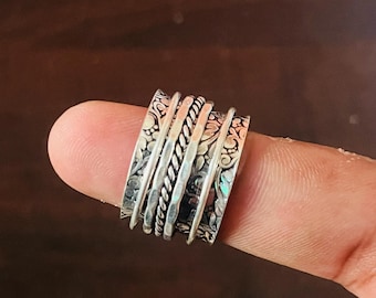 May include: A silver spinner ring with multiple bands. The bands feature intricate floral patterns and twisted rope designs. The ring is displayed on a finger, showcasing its details and craftsmanship.