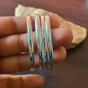 May include: A set of four silver-colored, thin, minimalist rings. The rings are stacked together, reflecting light. The background includes a light-colored stone and a dark brown surface.