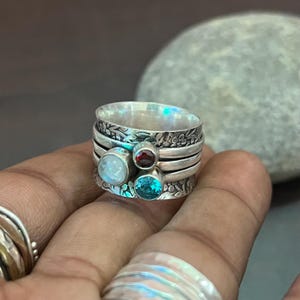 May include: A silver spinner ring with three gemstones: a light blue, a red, and a teal stone. The ring has floral engravings and multiple bands. The ring is held in a hand.