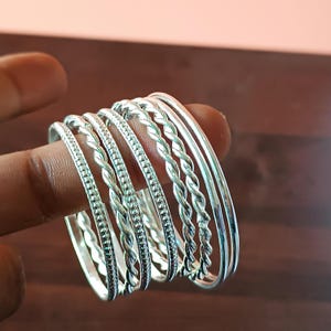 May include: A collection of silver bangles, featuring various designs. Some bangles have a braided pattern, while others have a simple, smooth surface. The bangles are displayed on a finger, showcasing their different textures and styles. The jewelry is made of a shiny, reflective metal.