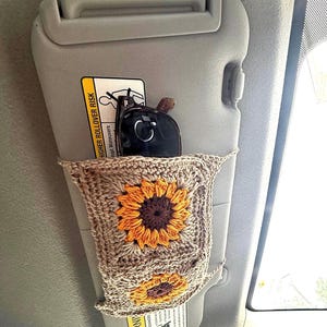 May include: A crocheted car visor organizer with a sunflower design. The organizer is tan with orange and brown sunflowers. Sunglasses and a key fob are stored in the pocket. The visor is gray.