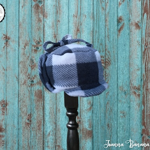 May include: A blue and white plaid fleece hat with a tie at the top. The hat is displayed on a black stand against a turquoise wooden background. The text "Joanna Banana Hats" is visible at the bottom of the image.